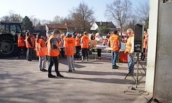 Eine Gruppe von Menschen in orangefarbenen Warnwesten steht in der Nähe eines Gebäudes. Ein Lautsprecher steht neben ihnen auf einem Ständer. Ein Traktor ist hinter ihnen geparkt.
