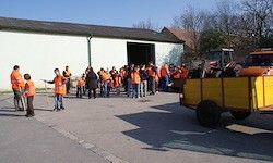 Eine Gruppe von Menschen in orangefarbenen Westen steht auf einem Parkplatz vor einem Gebäude. Ein gelber Anhänger ist in der Nähe geparkt.