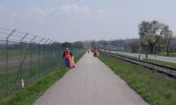 Zwei Personen in oranger Uniform laufen auf einem gepflasterten Weg neben einem eingezäunten Bereich und Gleisen.