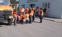 Eine Gruppe von Kindern und Erwachsenen in reflektierenden Westen versammelt sich neben einem Lastwagen vor einem Gebäude mit großen Fenstern.