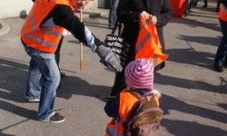 Ein Mann in Reflektorkleidung führt ein Kind mit Rucksack, beide halten orange Flaggen. Sie stehen auf einer Straße, mit Menschen und Gebäuden im Hintergrund.