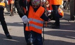 Ein Kind in einer orangefarbenen Warnweste hält einen Stock in der Hand, geführt von einem Erwachsenen, mit anderen Erwachsenen im Hintergrund, die Reinigungswerkzeuge halten.