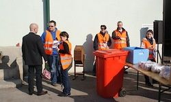 Eine Gruppe von Menschen in orangefarbenen Westen steht in der Nähe eines Tisches mit verschiedenen Behältern. Ein Mann im Anzug spricht mit einer Frau mit Rucksack.