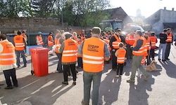 Eine Gruppe von Menschen in reflektierenden orangefarbenen Westen steht vor einem Gebäude, einige tragen Mützen. Ein roter Container ist neben ihnen platziert. Ein Baufahrzeug ist im Hintergrund geparkt.