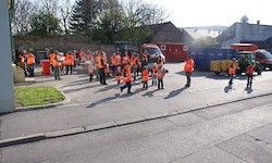 Eine Gruppe von Menschen in orangefarbenen Westen geht auf einer Straße, mit Fahrzeugen und Gebäuden im Hintergrund.