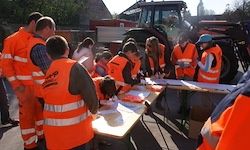 Eine Gruppe von Personen in orangefarbenen Westen steht um einen mit Papieren gefüllten Tisch herum. Sie stehen im Freien in der Nähe schwerer Maschinen.