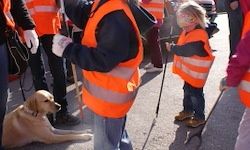 Zwei Kinder mit einem Blindenführhund tragen orange Westen und halten Stöcke. Sie stehen mit mehreren Erwachsenen, einige in orange Westen, auf einer Straße.