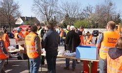 Eine Gruppe von Personen in orangefarbenen Warnwesten steht in einem Außenbereich mit Tischen und Kisten. Bäume und Autos sind im Hintergrund.