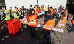 Eine Gruppe von Menschen in Sicherheitswesten steht um einen Tisch mit einem roten Mülleimer herum, möglicherweise bei einer Gemeinschaftsveranstaltung oder einer Aufräumaktion.