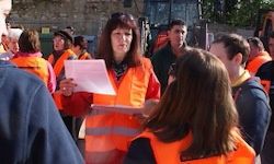 Eine Frau in einem orangefarbenen Sicherheitsweste hält ein Papier in der Hand, während sie mit einer Gruppe von Menschen spricht, die ähnliche Westen tragen, mit einem Gebäude und Grünpflanzen im Hintergrund.
