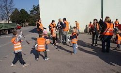 Eine Gruppe von Menschen in orangefarbenen Westen läuft auf einer Straße. Einige tragen Stöcke. Ein Lastwagen steht links. Bäume und ein Gebäude sind im Hintergrund.