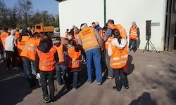 Eine Gruppe von Personen in orangefarbenen Warnwesten steht in einem Kreis vor einem Gebäude mit weißer Wand. Einige tragen Handschuhe und Mützen. In der Nähe steht ein Lautsprecher.