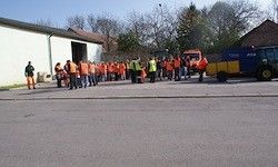 Eine Gruppe von Arbeitern, die Sicherheitswesten und orange Weste tragen, versammelt sich vor einem Gebäude. Ein gelbes und blaues Fahrzeug ist in der Nähe geparkt.