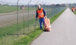 Eine Person in einem orangefarbenen Sicherheitsweste steht auf einem Bürgersteig, hält eine Tasche in der Hand und befindet sich in der Nähe eines Maschendrahtzauns.