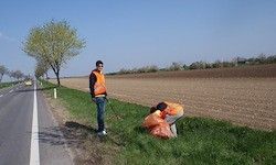 Ein Mann in einem orangefarbenen Vest steht neben der Straße, möglicherweise sammelt er Müll. Eine andere Person in einem orangefarbenen Vest ist auf dem Gras gebeugt.