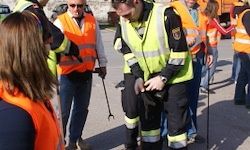 Eine Gruppe von Menschen in Warnkleidung trifft sich auf einer Straße. Ein Mann hält eine Harke, während ein anderer gebückt ist.