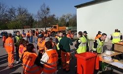 Eine Gruppe von Menschen in orangefarbenen Sicherheitsanzügen versammelt sich vor einem Gebäude mit einem gelben Lkw. Sie sind in der Nähe von Tischen mit Gegenständen und einem roten Container.