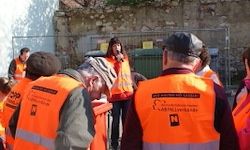Eine Gruppe von Personen in reflektierenden orangefarbenen Westen steht draußen, eine Frau spricht in ein Mikrofon. Dahinter befindet sich ein Metallzaun und ein Gebäude.