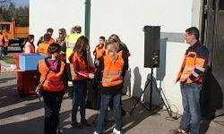 Eine Gruppe von Menschen in orangefarbenen Westen steht in der Nähe eines Gebäudes. Ein Lautsprecher auf einem Stativ ist aufgestellt. Ein Mann steht neben ihnen. Bäume sind im Hintergrund zu sehen.