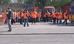 Eine Gruppe von Menschen in orangenen Westen steht auf einer Straße mit einem Maschinenfahrzeug im Hintergrund. Bäume und Gebäude sind in der Ferne sichtbar.