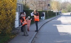 Eine Gruppe von Menschen in orangen Westen und Handschuhen sammelt Müll auf dem Bürgersteig. Einer macht Notizen.
