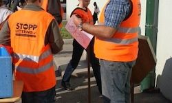 Eine Gruppe von Menschen in orange Westen steht neben einem Gebäude. Ein Mann hält eine Papierrolle und eine Dose und spricht mit einem anderen Mann. Ein blauer Kasten und ein Mülleimer stehen auf dem Boden.
