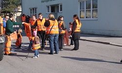 Eine Gruppe von Menschen in orangefarbenen Westen steht auf einer Straße, wahrscheinlich in Vorbereitung auf eine Gemeinschaftsaktion zur Müllsammlung. Im Hintergrund befindet sich ein Gebäude mit mehreren Fenstern.