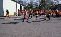 Eine Gruppe von Menschen in orangefarbenen Sicherheitswesten und Schuhen geht in einer Reihe auf einem gepflasterten Weg. Bäume und Gebäude sind im Hintergrund.