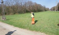 Eine Person in einer gelben Weste sammelt Müll in einem Grasfeld neben einer unbefestigten Straße. Die Bäume sind kahl, und der Himmel ist klar.