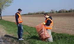 Zwei Männer in Warnwesten sammeln Müll am Straßenrand, mit einem weiten Feld dahinter unter klarem Himmel.