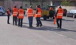 Eine Gruppe von Menschen in orangefarbenen Westen geht auf einer Straße. Im Hintergrund befinden sich Gebäude, geparkte Autos und ein gelber Traktor.