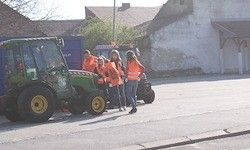 Eine Gruppe von Menschen in orange Westen steht neben einem grünen Traktor auf einer Straße. Ein blauer Lkw ist hinter ihnen geparkt. Gebäude und Bäume sind im Hintergrund zu sehen.