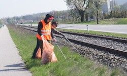 Zwei Personen in orangenen Westen räumen Müll in einem Grasbereich neben Gleisen, einer Straße und Bäumen auf.