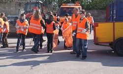 Eine Gruppe von Freiwilligen in reflektierenden orangefarbenen Westen läuft auf einer Straße, mit Maschinen und einem gelben Lastwagen im Hintergrund.