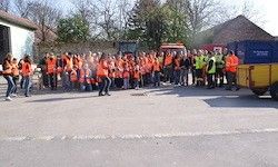 Eine Gruppe von Menschen in hochsichtbaren orangefarbenen Westen und Hosen steht auf der Straße, möglicherweise an einer Gemeinschaftsveranstaltung teilnehmend, mit Gebäuden, Bäumen und Maschinenfahrzeugen im Hintergrund.