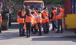 Eine Gruppe von Menschen in reflektierenden orangefarbenen Westen steht auf einem Parkplatz mit Baufahrzeugen im Hintergrund. Einige halten weiße Flaggen.