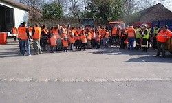 Eine Gruppe von Menschen in Warnwesten steht auf der Straße und blickt auf ein Gebäude und Fahrzeuge. Im Hintergrund befinden sich Bäume und Häuser.