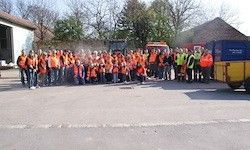 Eine Gruppe von Menschen in orangefarbenen Westen steht vor einem gelben und blauen Lastwagen auf einer asphaltierten Straße. Dahinter stehen mehrere Bäume und ein Gebäude mit einem Dach.