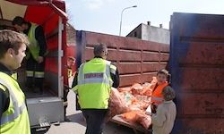 Eine Gruppe von Menschen in Sicherheitswesten lädt Säcke in einen großen Müllcontainer. Einige Säcke sind bereits drin. Sie stehen vor einem Gebäude mit einer Straßenlaterne.