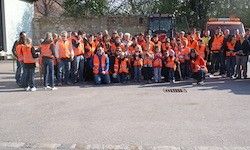Eine große Gruppe von Menschen in orangenen Westen posiert für ein Foto im Freien, mit Gebäuden und Fahrzeugen im Hintergrund.