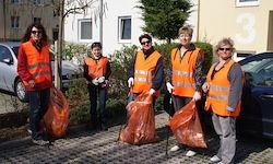 Fünf Personen in orange Westen und Handschuhen halten orange Mülltüten in einem gepflasterten Bereich. Ein Gebäude und Autos sind im Hintergrund.