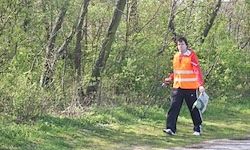 Eine Person in einer orangefarbenen Weste geht auf einem grasigen Pfad, umgeben von Bäumen und Büschen.