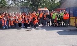 Eine Gruppe von Personen in Warnwesten posiert für ein Foto auf einer Straße, mit Maschinenfahrzeugen im Hintergrund.