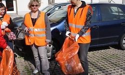 Vier Personen in orange Warnwesten, die orange Plastiktüten halten, stehen neben einem blauen Auto. Eine Person hält einen Stock.