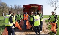 Eine Gruppe von Menschen in Warnwesten steht um einen roten Anhänger herum und hält orangefarbene Tüten in der Hand, wahrscheinlich um einen Feldweg zu reinigen.