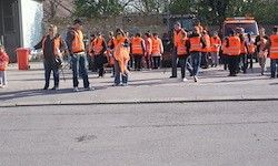 Eine Gruppe von Menschen in orangefarbenen Westen steht in einer Reihe, möglicherweise auf Anweisungen wartend. Einige tragen Ausrüstung. Ein Gebäude und ein roter Mülleimer sind in der Nähe.