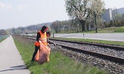 Zwei Personen in orange Weste sammeln Müll entlang einer Bahnlinie ein, mit einer Straße und Grünflächen in der Nähe.