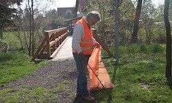 Eine Person in einem orangefarbenen Weste hält einen Müllbeutel und sammelt Müll in der Nähe einer Holzbrücke in einem grünen Bereich mit Bäumen.