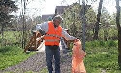 Ein Mann in einem orangefarbenen Sicherheitsweste sammelt Müll auf einem Kiesweg. Der Weg ist von Bäumen und Gras umgeben. Im Hintergrund befindet sich eine Holzbrücke.