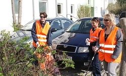 Drei Personen in orangefarbenen Westen stehen neben einem blauen Auto und scheinen einen Garten zu reinigen oder zu pflegen. Hinter ihnen sind mehrere Autos vor einem Gebäude geparkt.
